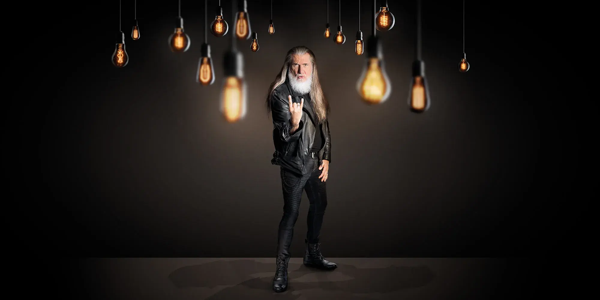 Bärtiger langhaariger Mann im Rocker-Outfit mit Metal-Geste in einem dunklen Raum mit mehreren herabhängenden Glühbirnen